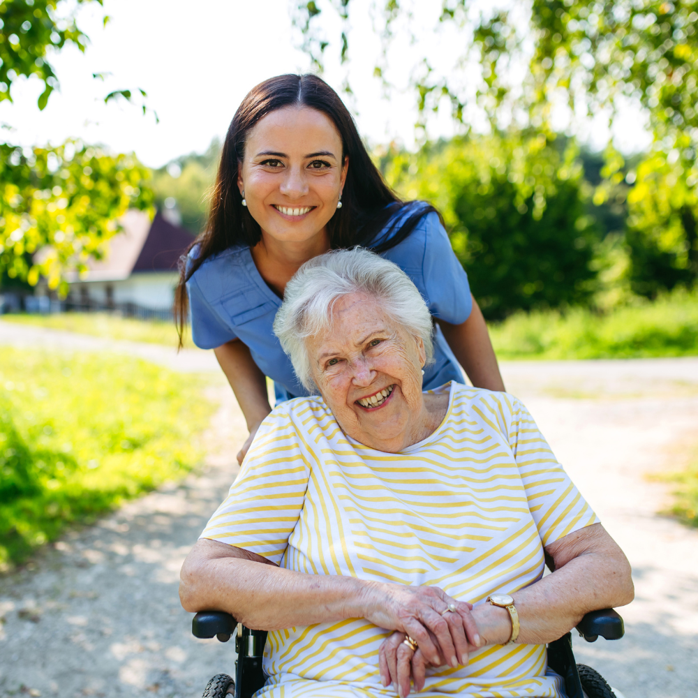 Une aidante souriante en tenue bleue se tient derrière une femme âgée assise dans un fauteuil roulant en extérieur, illustrant l'accompagnement et la mobilité retrouvée.