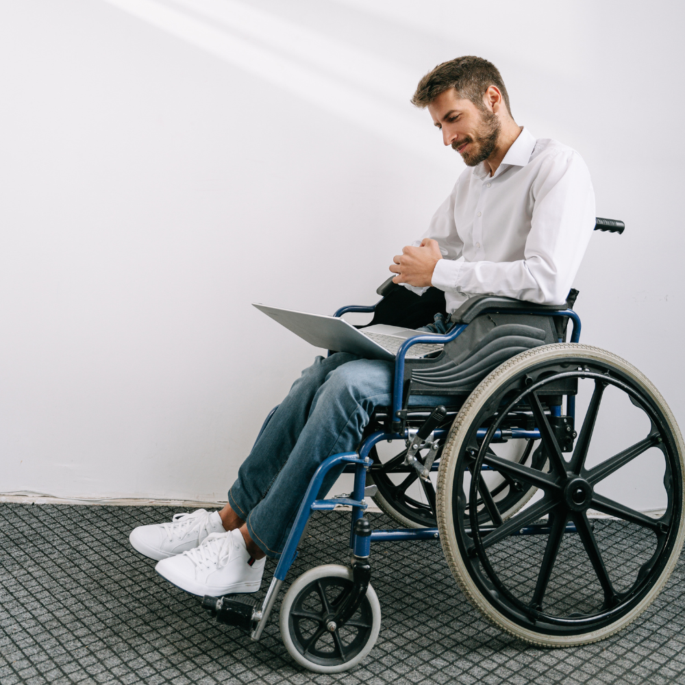Un homme assis dans un fauteuil roulant manuel utilise un ordinateur portable sur ses genoux devant un mur blanc, symbolisant l'autonomie et l'intégration facilitée par les nouveaux équipements.