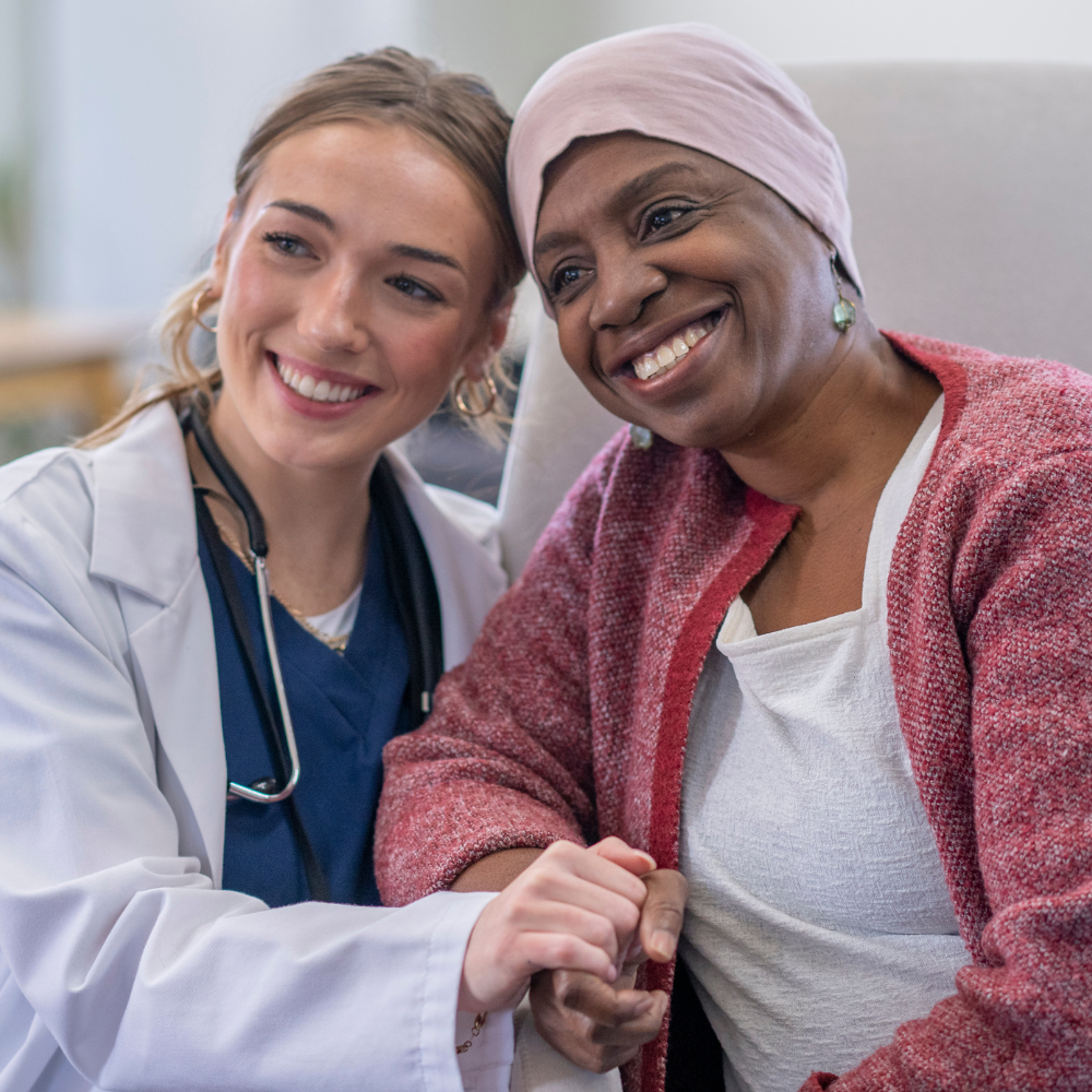 Une jeune professionnelle de santé en blouse blanche sourit avec empathie en tenant la main d’une patiente portant un turban rose, illustrant la solidarité et l'accompagnement humain face au cancer.